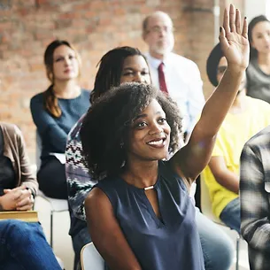 Image of a seated woman holding hand up in amongst other seated people listening to a presentation